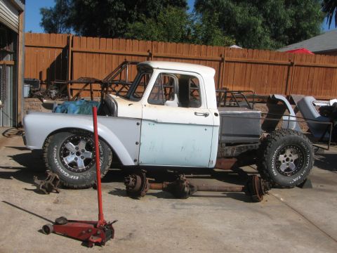 1990 Ford Bronco - vintage two-tone pickup body sits on axle stands with bare axles and a floor jack during a chassis build in a fenced yard.