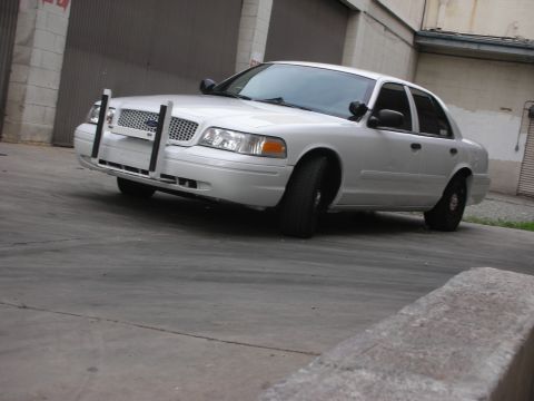 2004 Ford Crown Victoria - A white Ford Crown Victoria with a push bumper is parked outside a building.