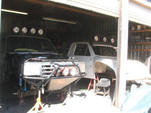 1990 Ford Bronco - Two off-road truck projects with roof light bars sit inside an open garage during assembly.