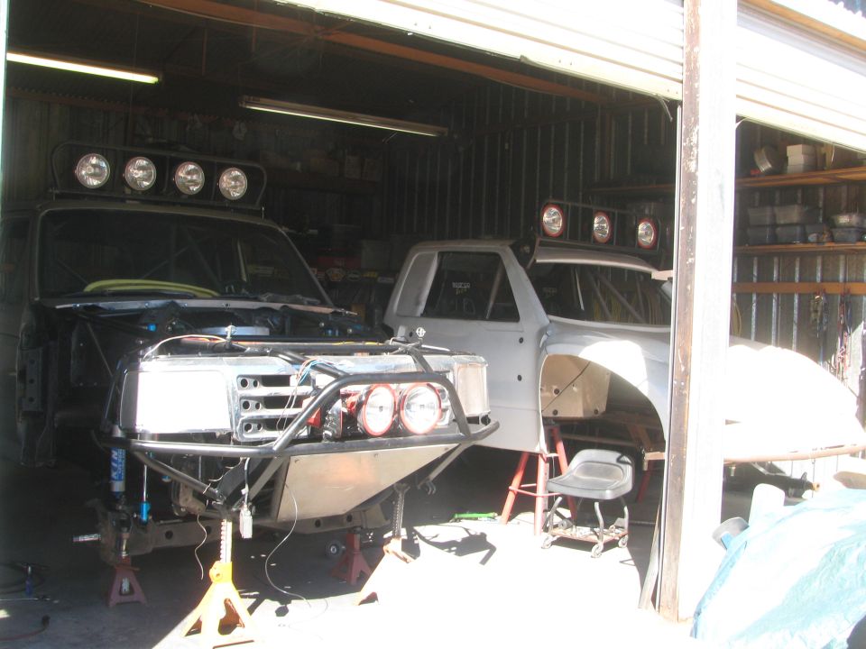 Two off-road truck builds parked inside a garage with overhead light bars