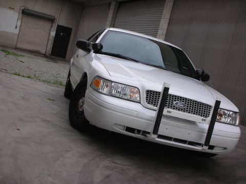 2004 Ford Crown Victoria - A white Ford Crown Victoria with a push bar is parked in front of a corrugated metal garage door.