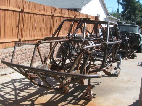 1990 Ford Bronco - bare welded tube-frame off-road chassis sits on jack stands in a driveway beside a wood fence.