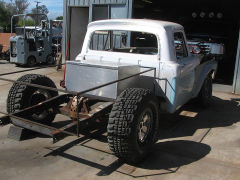 1990 Ford Bronco - Rear view of a white vintage pickup cab mounted on a bare frame with a spare tire, parked in mid-build by a garage.