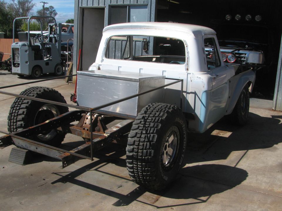 Rear of vintage pickup cab on a bare frame with spare tire, mid-build by a garage