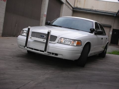 2004 Ford Crown Victoria - A white Ford Crown Victoria, possibly a police car, is parked in front of a garage with a push bumper attached.