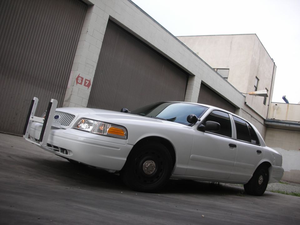 White 2004 Ford Crown Victoria police car parked in front of a garage. It has a push bumper and black steel wheels