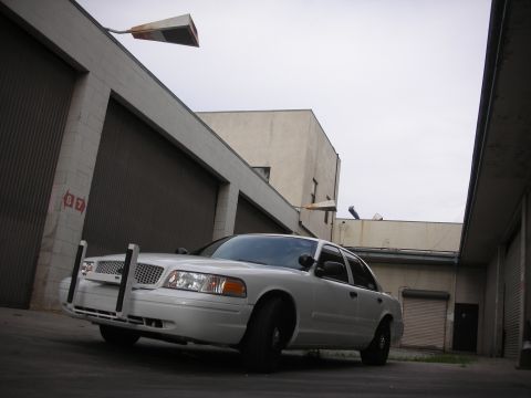 2004 Ford Crown Victoria - A white 2004 Ford Crown Victoria with a push bar is parked in an alley between buildings.