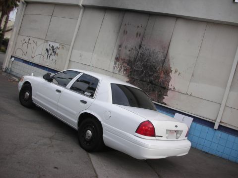 2004 Ford Crown Victoria - A white 2004 Ford Crown Victoria is seen from the rear-three-quarter angle, parked beside a weathered wall.