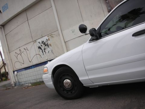 2004 Ford Crown Victoria - Side profile of a white sedan with a black side mirror, parked on asphalt near a textured wall with graffiti.
