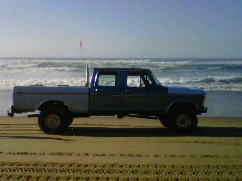 1976 Ford F-250 - A blue and gray Ford F-Series truck sits on the sand near the ocean with waves crashing.