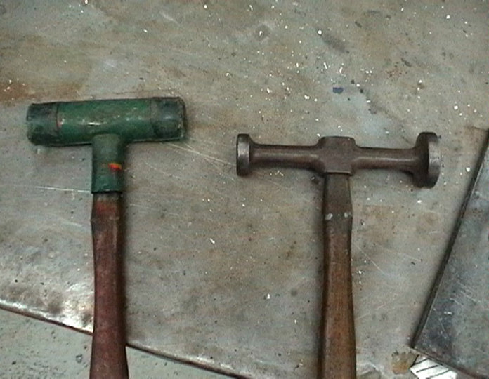 Two body hammers on a workbench. One has a green head and wooden handle, the other has a metal head and wooden handle