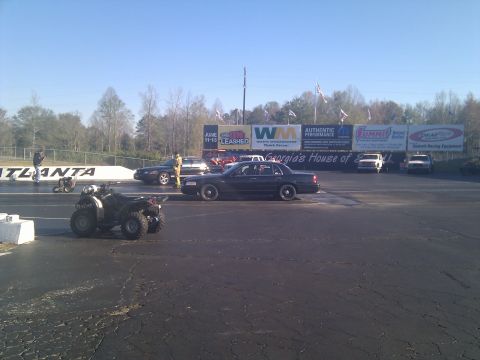 2001 Ford Crown Victoria - A dark 2001 Ford Crown Victoria is parked on a drag strip, with an ATV in the foreground and advertising banners in the background.