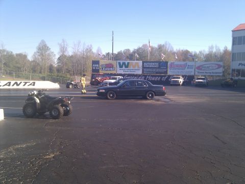 2001 Ford Crown Victoria - A police car sits on the drag strip with ATVs and other vehicles in the background under a clear sky.