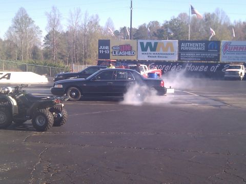 2001 Ford Crown Victoria - A black 2001 Ford Crown Victoria performs a burnout on a drag strip, with other vehicles and advertising in the background.