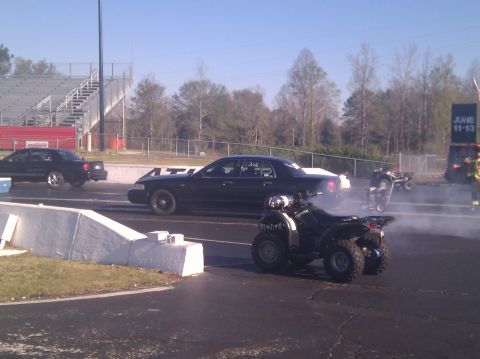 2001 Ford Crown Victoria - A black 2001 Ford Crown Victoria sedan is seen at a drag strip alongside ATVs, with smoke billowing from one of the ATVs.