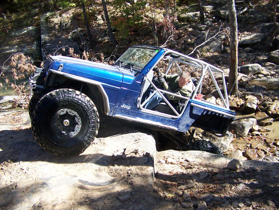 Blue Jeep Wrangler with roll cage off-roading over rocks and mud