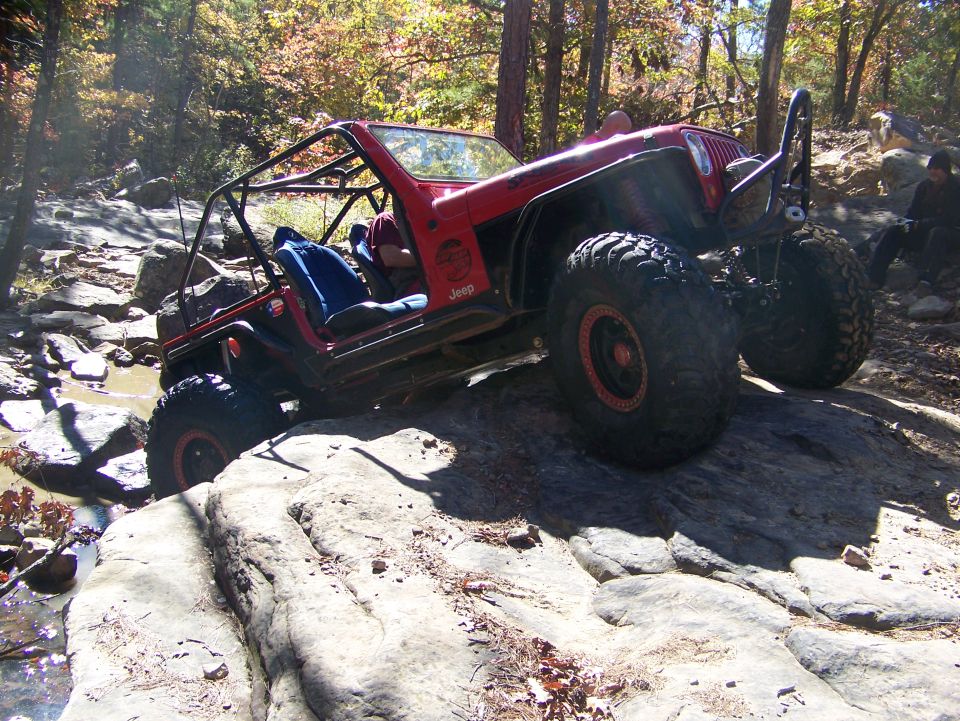 Red Jeep Wrangler with roll cage and large tires navigating rocky terrain