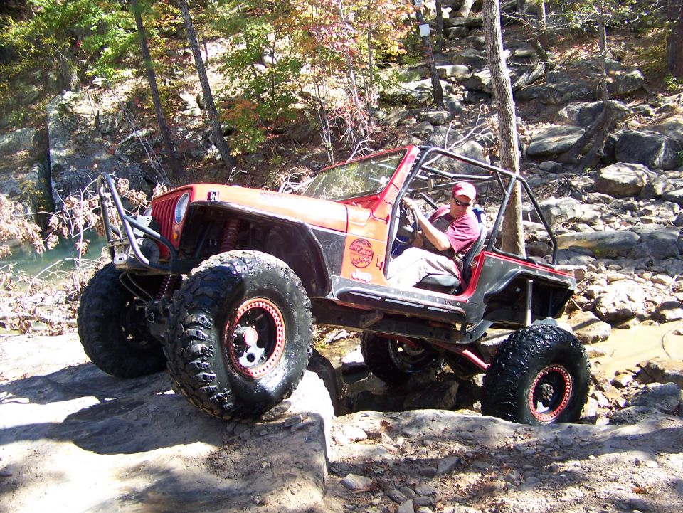 Red Jeep Wrangler with large tires navigating rocky terrain. Driver wearing a red cap and shirt