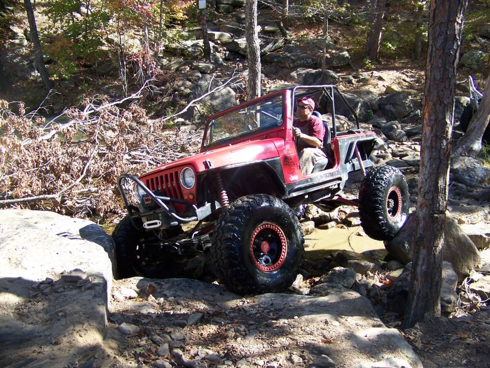 Red Jeep Wrangler with large tires navigating rocky terrain, driver visible