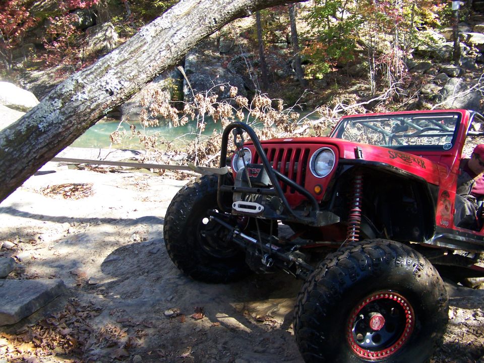 Red Jeep Wrangler modified for off-roading, parked on a rocky trail near a river