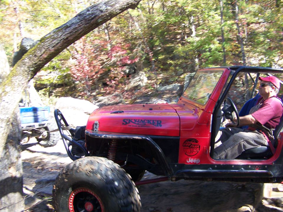 Red Jeep Wrangler with "SKYJACKER" decal on hood, driving on rocky terrain. Another vehicle is visible in the background