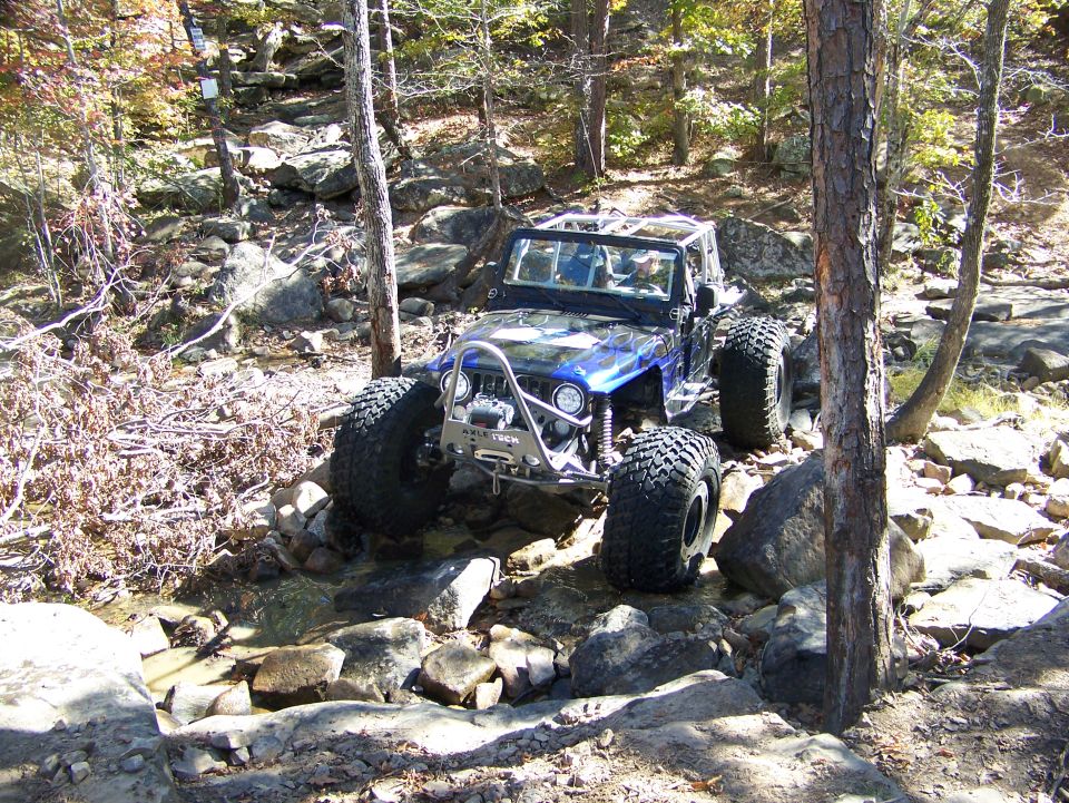 Blue flamed caged Jeep rock crawler climbing rocky creek bed in fall woods