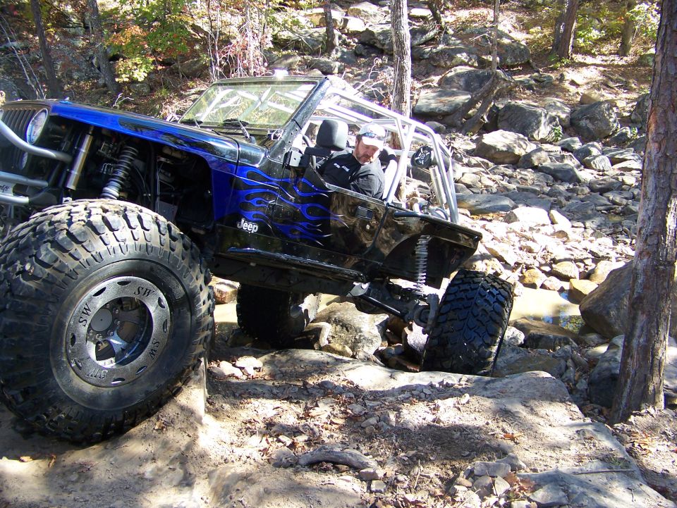 Modified Jeep Wrangler with large tires navigating rocky terrain. Blue flame decals on the side