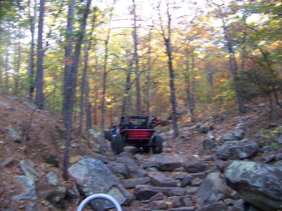 Red off-road vehicle navigating a rocky trail in a forest with autumn foliage