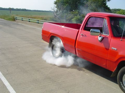 1986 Ford Bronco - A red pickup truck is performing a burnout on a paved road, creating a cloud of smoke from its rear tire.