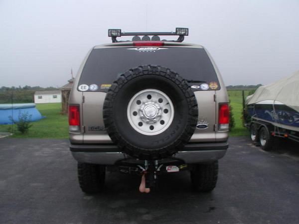 Rear of a tan Ford Excursion SUV lifted with rear spare tire, roof light bar and trailer hitch in a rural driveway