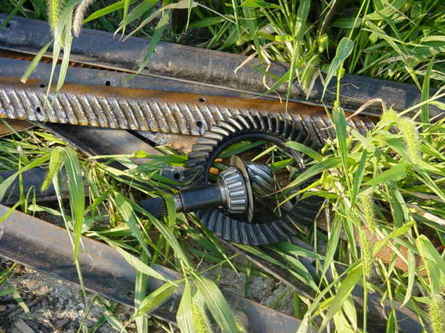 Ring-and-pinion gear set lying in tall grass on a steel beam, pinion bearing visible