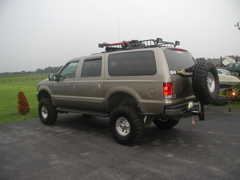 Lifted tan Ford Excursion SUV with big tires, roof rack and spare, rear 3/4 view on driveway