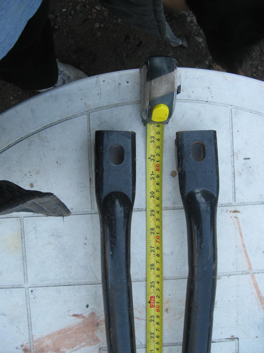 Two black metal parts with holes, measured by a yellow tape measure