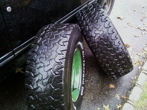 1990 Ford Bronco - Two off-road tires are shown, one with a green rim, leaning against a black vehicle on a wet surface.