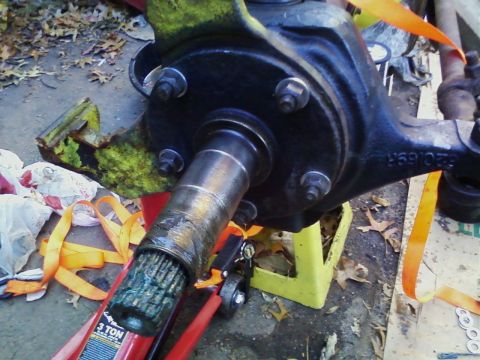 1990 Ford Bronco - Close-up of a bare front differential housing propped on a yellow jack stand and red floor jack on a leaf-strewn driveway, with the splined…