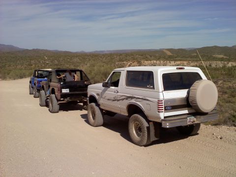 1996 Ford Bronco - A convoy of SUVs, including a white 1996 Ford Bronco, travels down a dusty desert road.