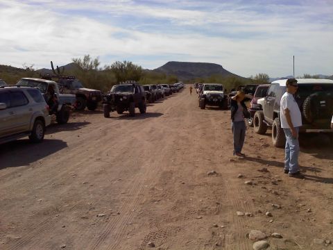1996 Ford Bronco - A convoy of off-road vehicles is parked on a dusty road, with a mountain in the background under a cloudy sky.