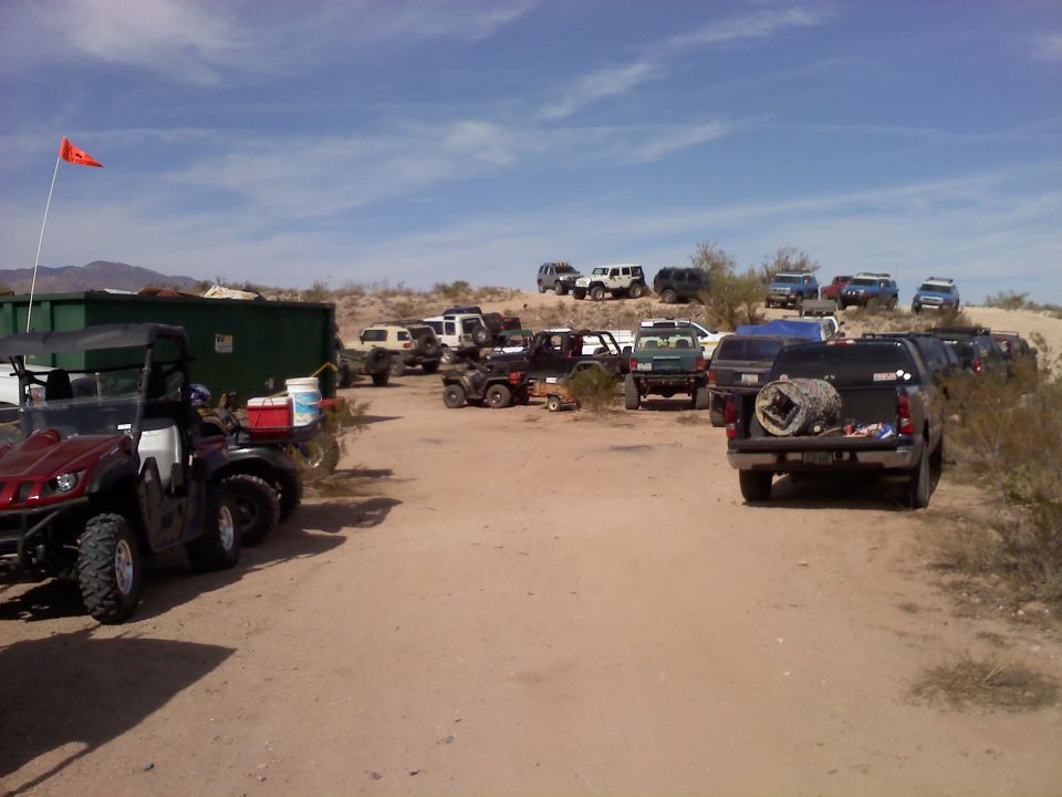 Desert scene with many parked SUVs, trucks, and ATVs under a blue sky with scattered clouds