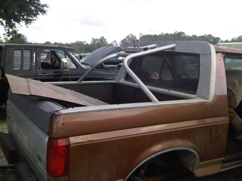 1992 Ford Bronco - A brown Ford Bronco with a roll cage and diamond plate trim is parked in a junkyard with other vehicles.