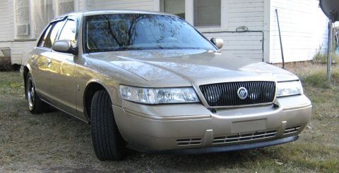 2000 Mercury Grand Marquis - A gold Mercury Grand Marquis is parked in front of a white house with trees reflected in the windshield.