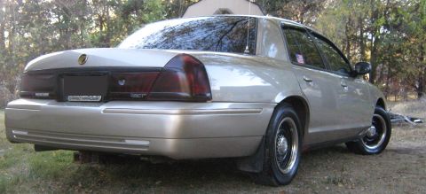 2000 Mercury Grand Marquis - Rear view of a tan 2000 Mercury Grand Marquis sedan parked on a dirt lot with trees in the background.