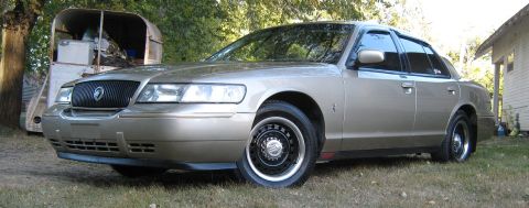 2000 Mercury Grand Marquis - A gold 2000 Mercury Grand Marquis sedan is parked on a grassy lawn next to a trailer and a house.
