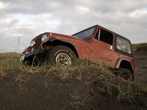 1996 Ford Bronco - A red Jeep CJ-7 is partially submerged in dark sand, with tall grass growing around it under a cloudy sky.