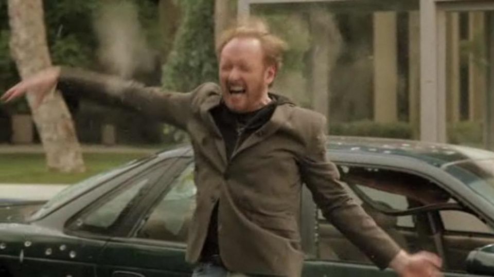 man in jacket flailing arms, car with bullet holes visible