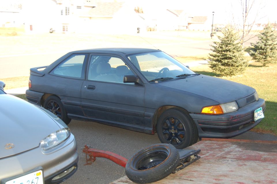 Dark gray 1991 Ford Escort hatchback parked next to a silver car, with a spare tire and red trailer hitch in the foreground