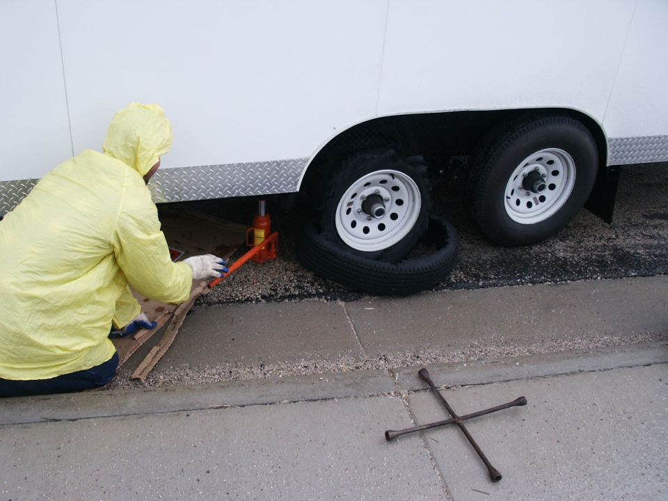 Person in yellow rain gear changing a flat tire on a white trailer with a jack and lug wrench