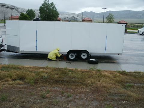 1990 Ford Bronco - A person in a yellow raincoat works on a flat tire of a white enclosed trailer on a wet day.