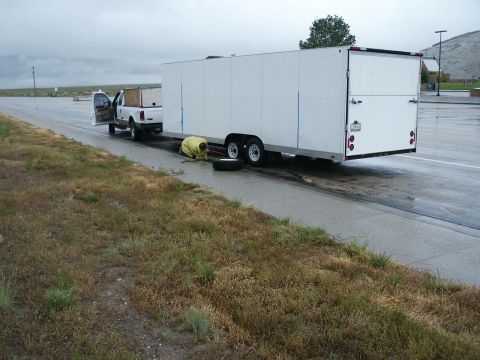 1990 Ford Bronco - A person changes a tire on a white cargo trailer being towed by a white pickup truck on a roadside.