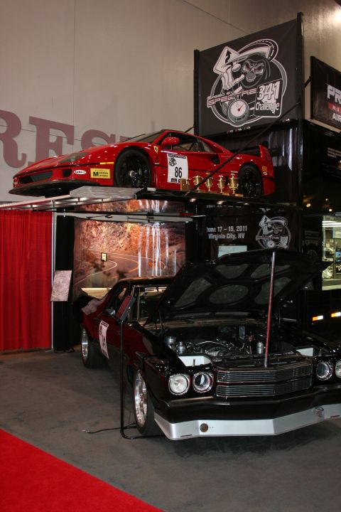 1998 Ford Crown Victoria - red mid-engine sports car sits on an elevated display rack above a black muscle car with its hood up at an indoor automotive trade show boo…