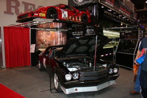 1998 Ford Crown Victoria - glossy black muscle car with its hood raised sits beneath a red sports car mounted on an overhead display rack at a busy indoor car show.